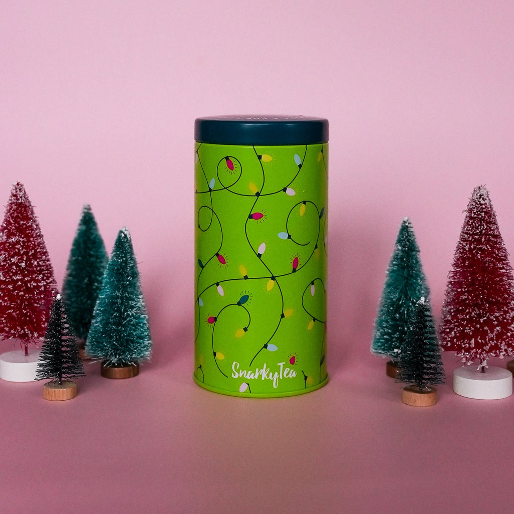 A cylindrical green tea tin with a dark teal lid, decorated with a pattern of colorful Christmas string lights on a bright lime green background. The brand name 'Snarky Tea' is printed in white script near the bottom.