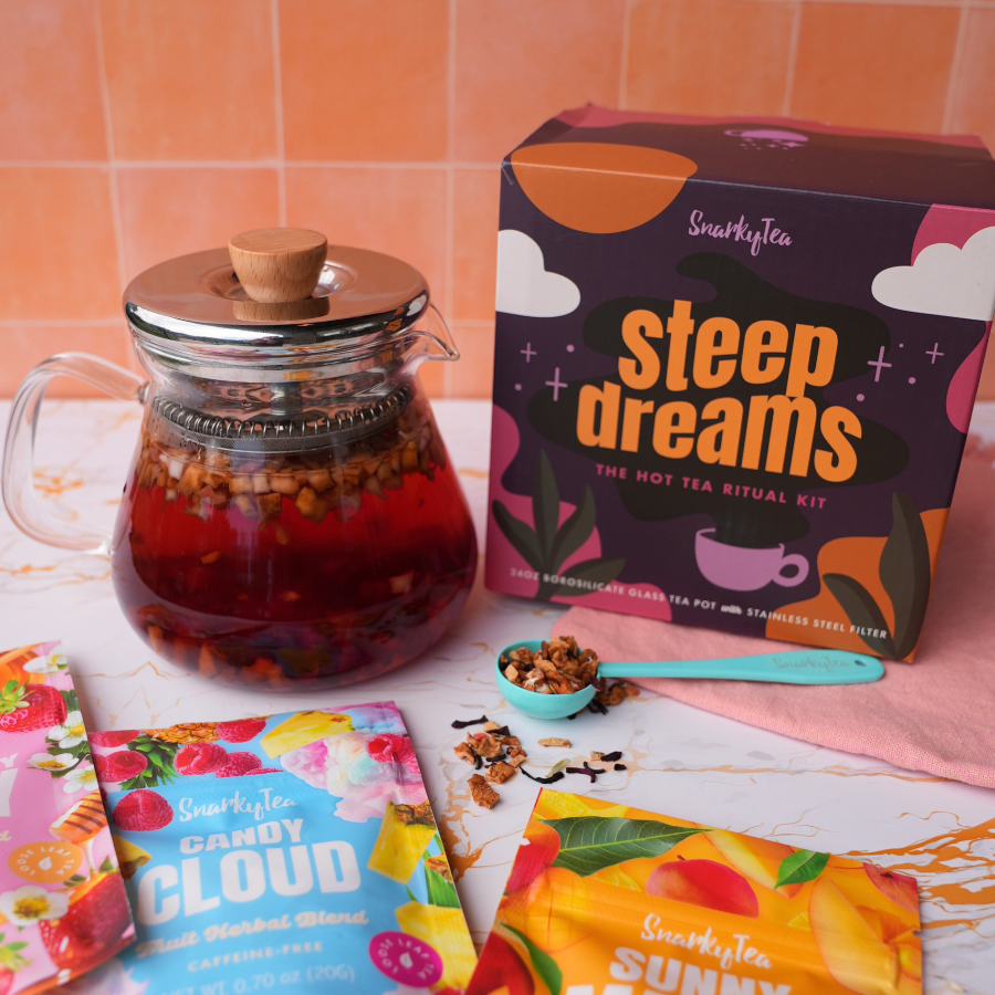 A clear glass teapot with a cork lid and metal infuser, shown next to its colorful retail box labeled 'steep dreams' by SmaraphyTea on a white background.