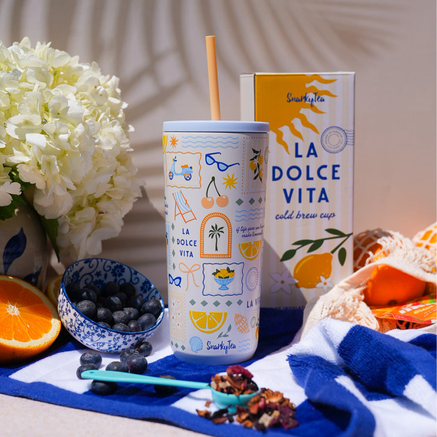 A whitebox photo of the la dolce vita cold brew cup next to the la dolce vita box. The cup is cream colored with an orange straw and blue lid with illustrations of lemons, seashells, sunglasses, and other summer-related items. The box is cream and yellow striped with a large sun and lemon with flowers next to it
