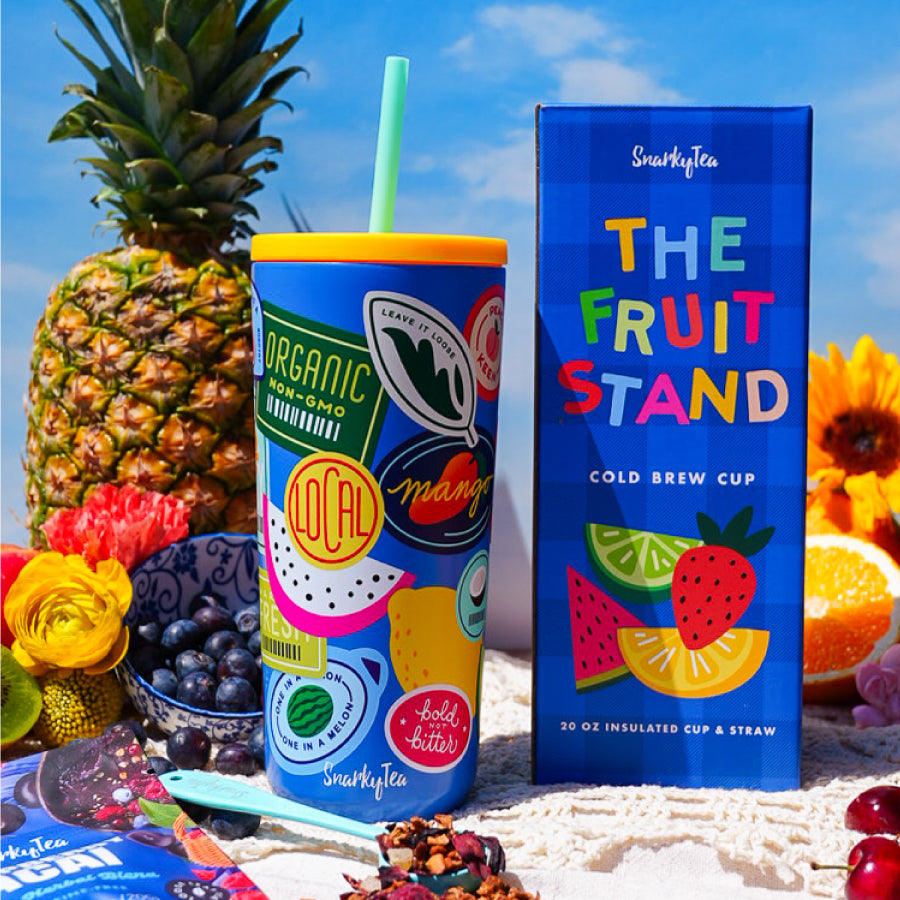 A whitebox image of the fruit stand cup next to the fruit stand box. The cup is a bright blue color with a teal straw and yellow lid with different fruit and sticker illustrations. The box is blue checkered with multicolored fonts and fruit illustrations on the front.  