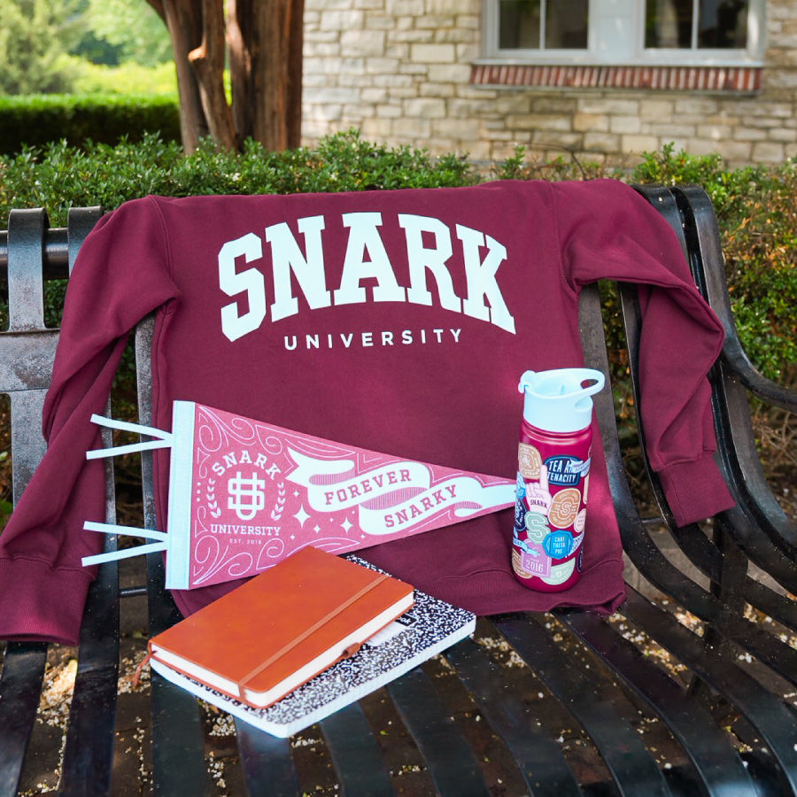 A woman wearing a maroon long-sleeve sweatshirt with 'SNARK UNIVERSITY' printed in white and light blue on the front, posed against a solid white background.